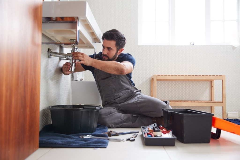 Person sitting on the floor under a sink wearing overalls with tools on the floor fixing a pipe